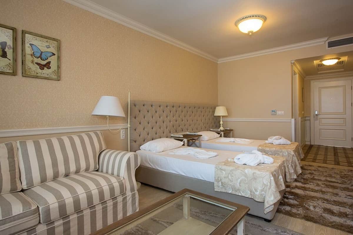 A hotel room with a striped sofa, a bed with two pillows, a bedside table with a lamp, and a small table with a glass top.
