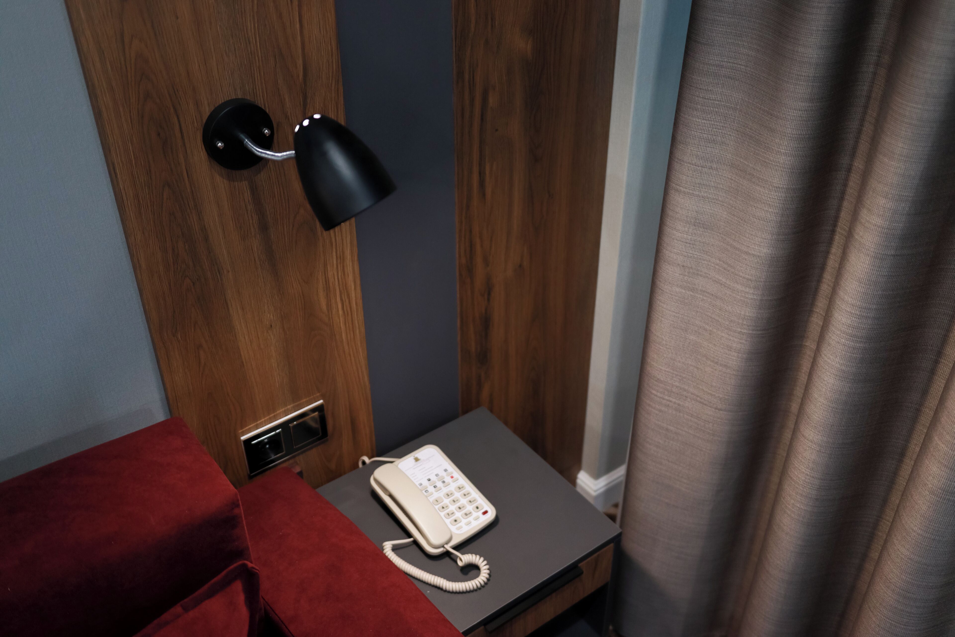 A hotel room with a wooden door, a red upholstered bench, a bedside table with a white telephone, and a black wall-mounted lamp.