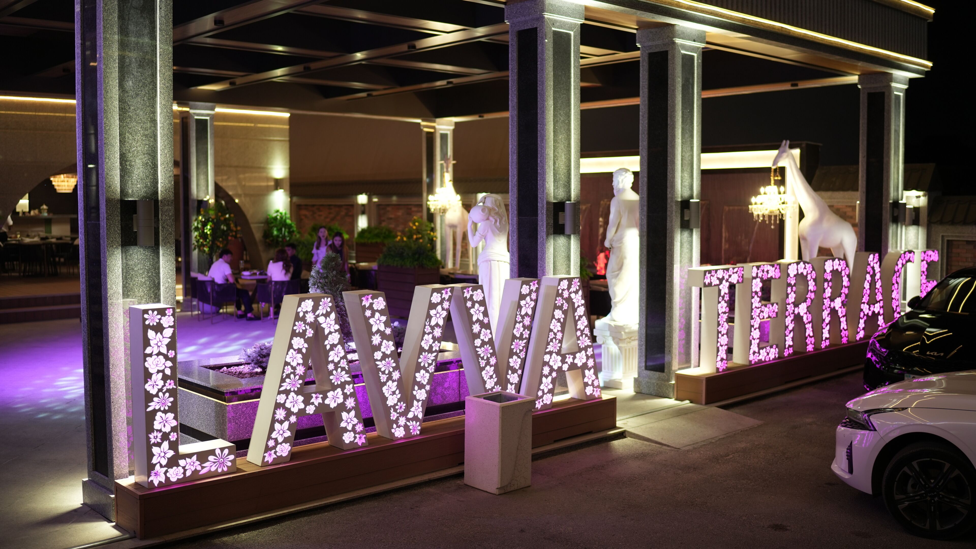 Entrada de hotel con letras iluminadas que dicen 'LAVARA TERRACE' y un automóvil blanco estacionado cerca.