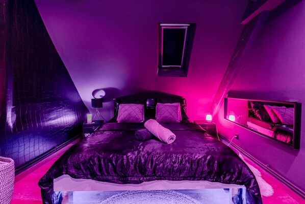 A bedroom with a black leather bed, a small bedside table, a nightstand, a wall-mounted TV, and a window with a black frame. - Love Room Alsace Escale du Désir (Niederbronn-les-Bains)