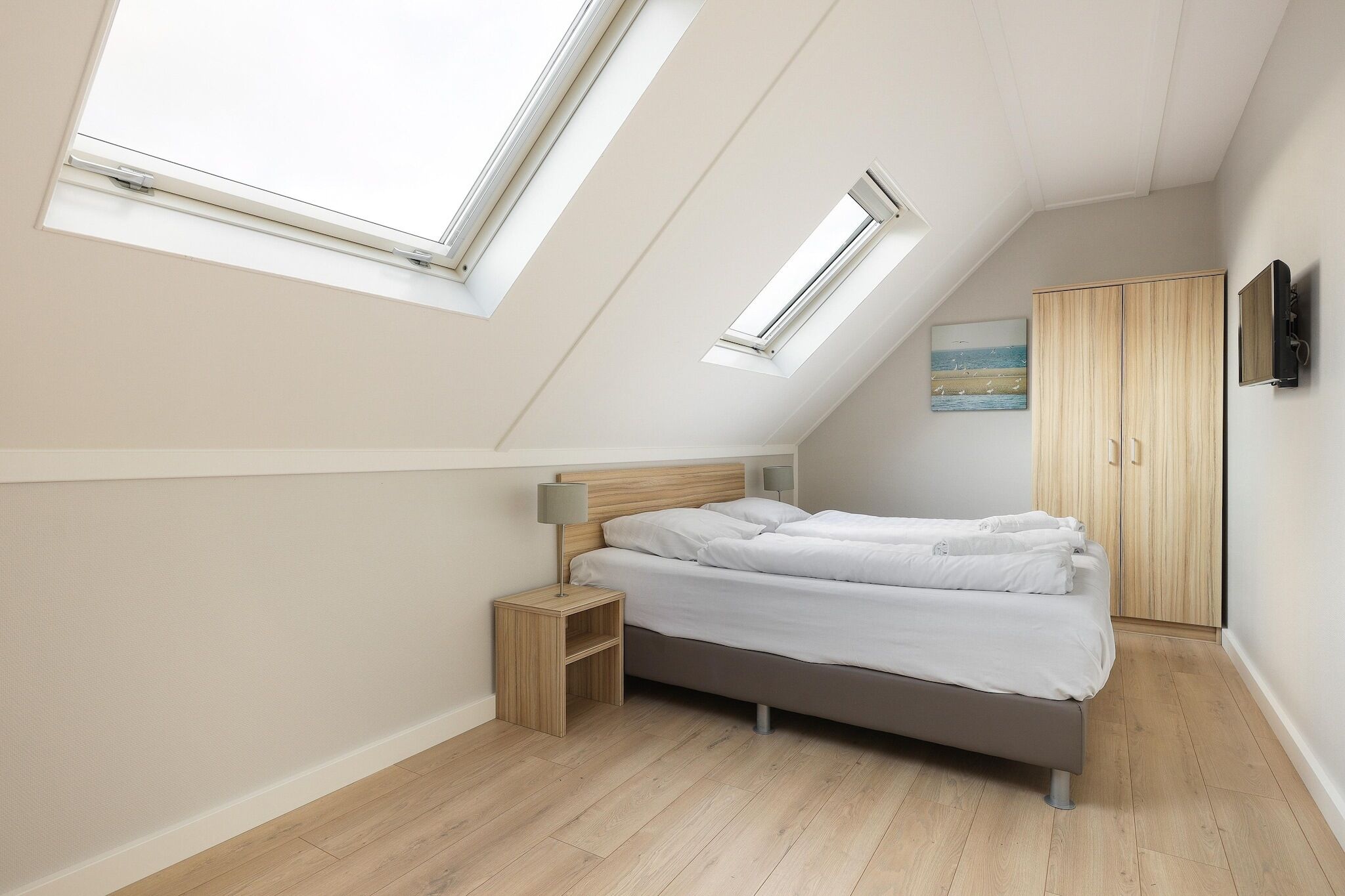 A bedroom with a sloped ceiling, skylights, a bed, a nightstand, a wardrobe, and a painting on the wall.