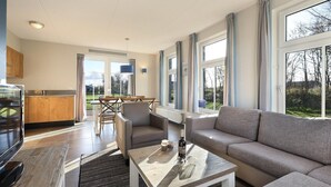 A modern living room with a sofa, a coffee table, and a TV. There is a kitchen area with a counter and cabinets. Large windows provide natural light and views of the outdoors. - Beach Retreat w/ Garden (Nieuwvliet)