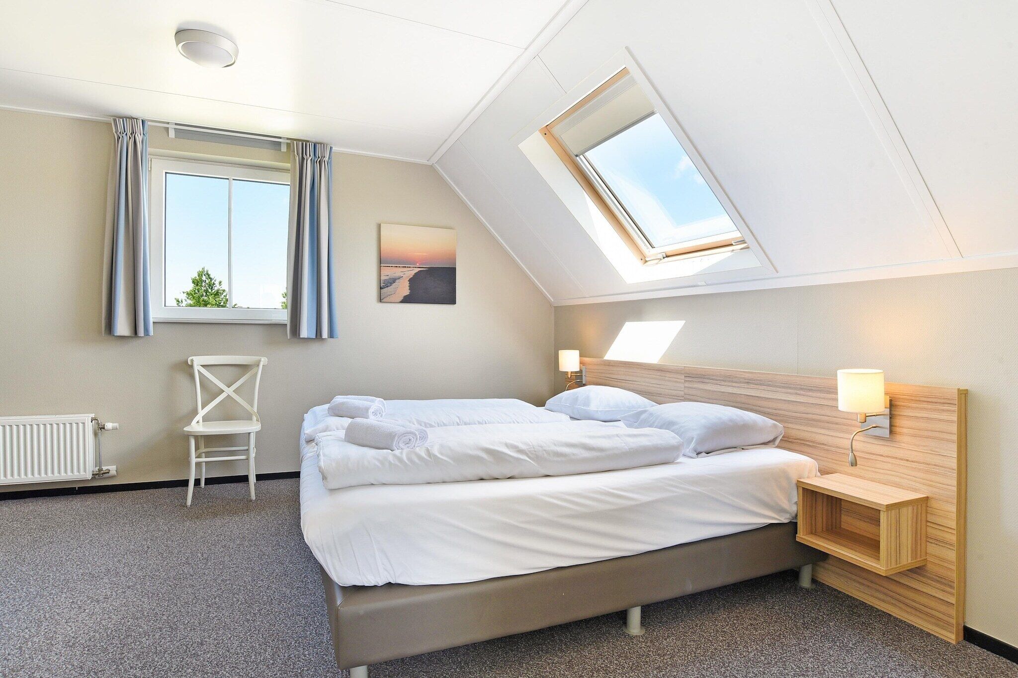 Een net opgemaakt bed met witte lakens, een nachtkastje met een lamp, een stoel en een dakraam boven het bed.