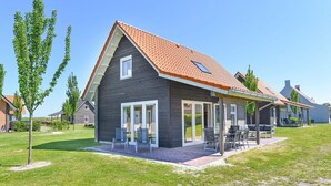 Une maison moderne à deux étages, dotée d’un toit recouvert de tuiles rouges, de grandes fenêtres et d’un patio couvert aménagé avec du mobilier d’extérieur.