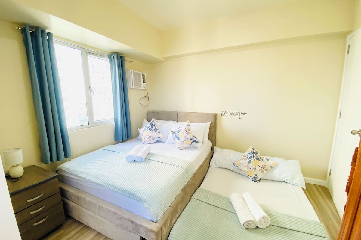 Habitación de hotel con dos camas, una ventana con cortinas, una mesita de noche con lámpara y un letrero decorativo en la pared.