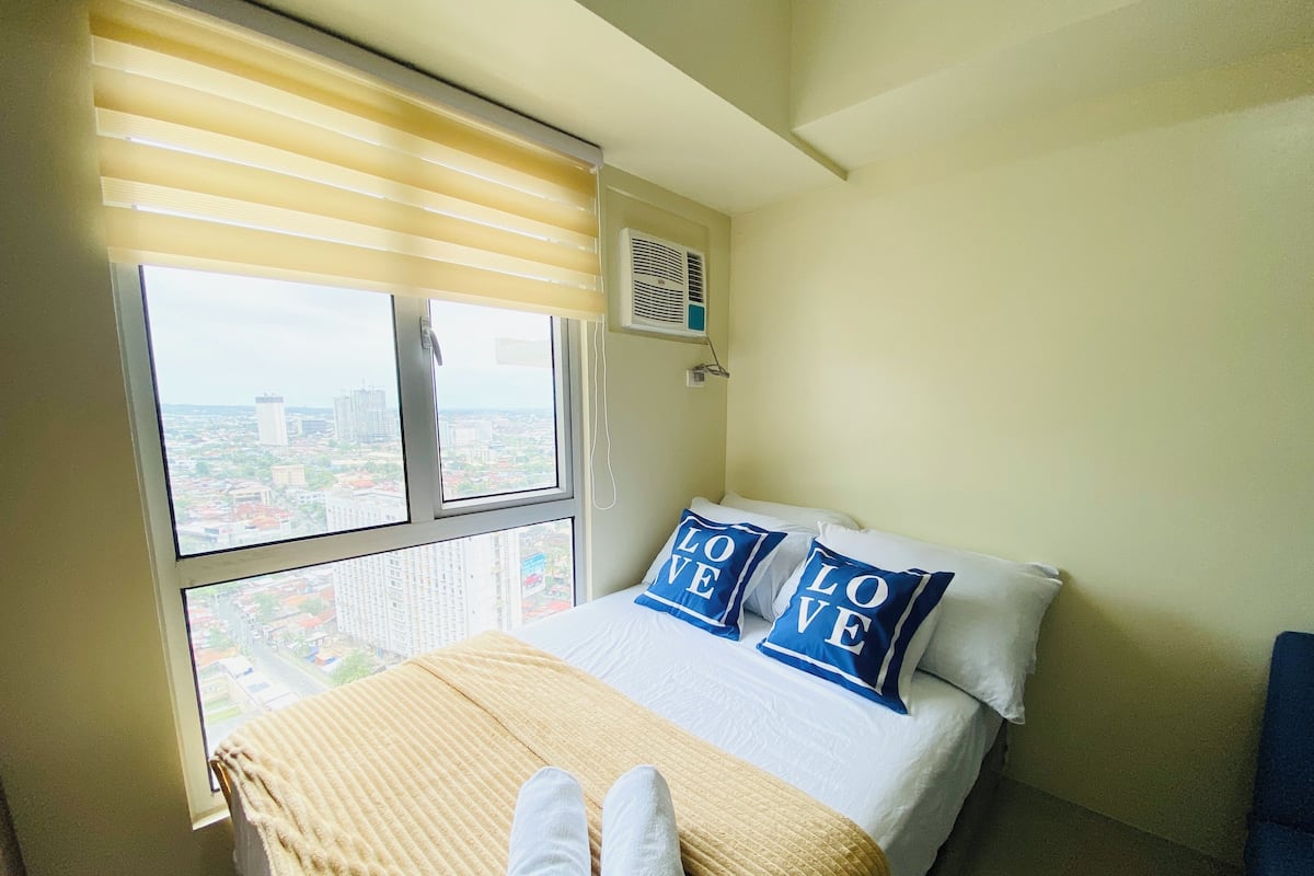 Un dormitorio con cama, dos almohadas con el texto "LOVE" y una ventana con persianas que dan a una vista de la ciudad.