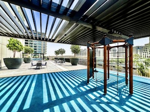 An outdoor area with a blue rubberized floor, large planters with trees, a wooden play structure, and a view of a cityscape with buildings in the background.