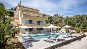 A modern two-story building with a swimming pool, sun loungers, and umbrellas.