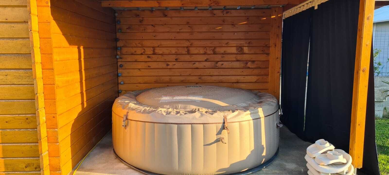 Un bain à remous dans un chalet en bois, avec une porte noire et une paire de pantoufles posées sur le sol.