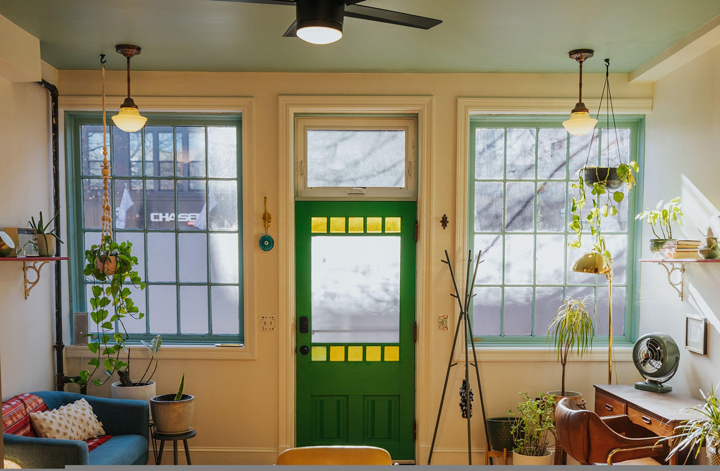 A cozy living room with a green door, two windows with plants, a ceiling fan, and a desk with a lamp.