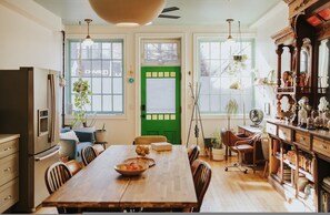 A dining area with a wooden table, chairs, and a green door. There are plants, a fan, and a bookshelf with various items. - Wyatt's Saloon Guest House 2 (Baltimore)