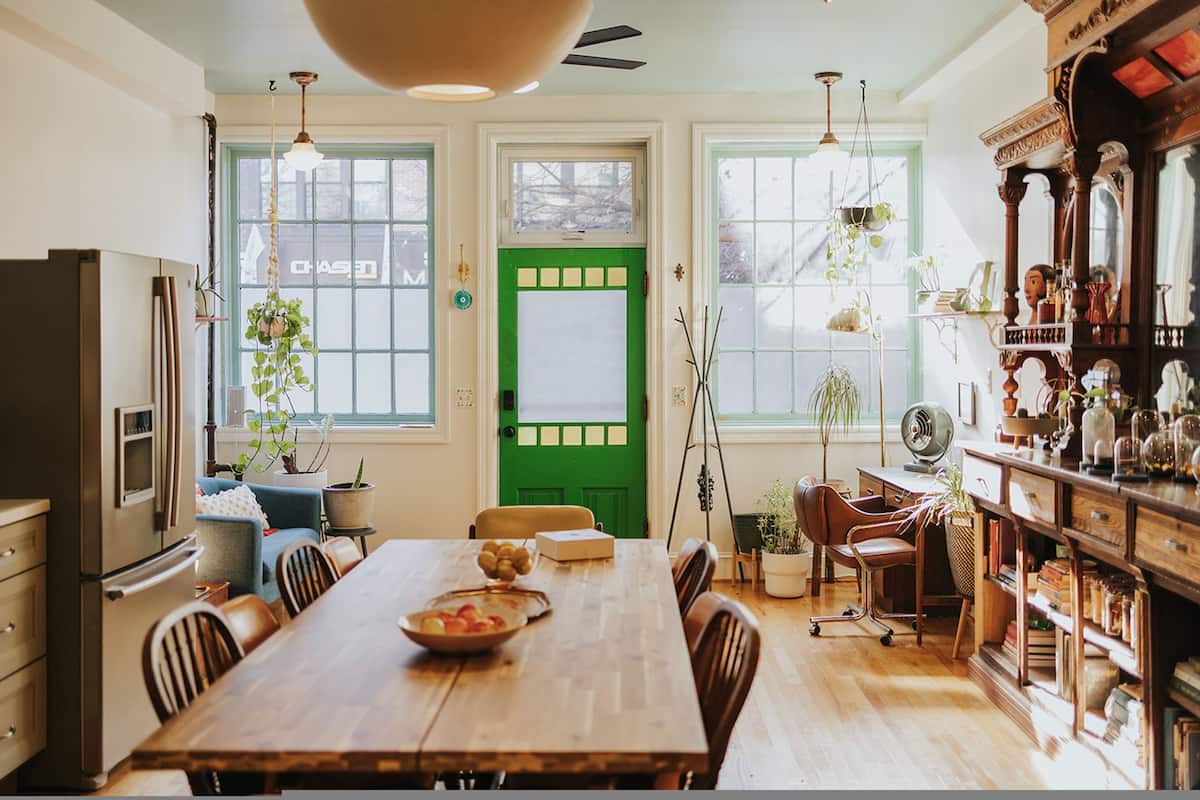 A dining area with a wooden table, chairs, and a green door. There are plants, a fan, and a bookshelf with various items.