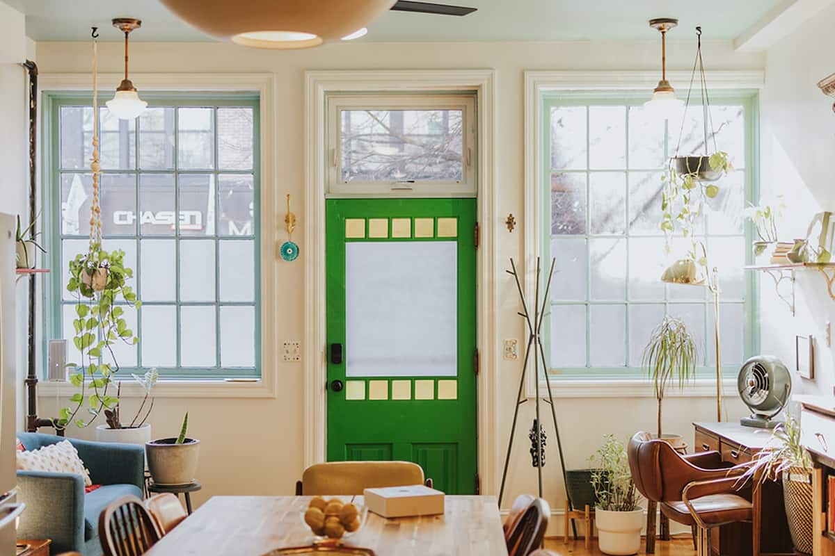 A dining area with a wooden table, chairs, and a bowl of fruit. There are potted plants and a green door with a frosted window.