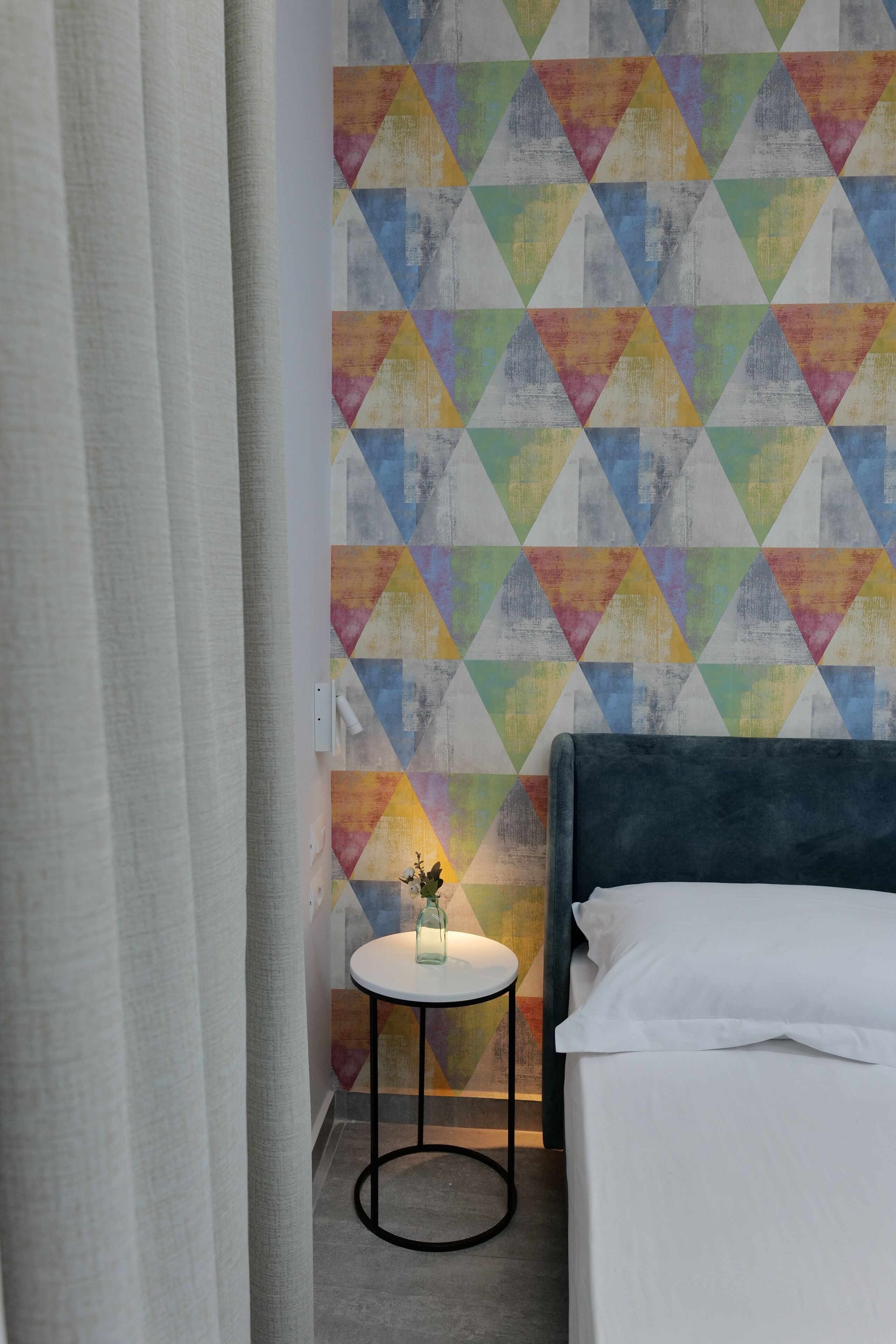 A hotel room with a bed, a small round table with a vase, and a colorful geometric wall design.