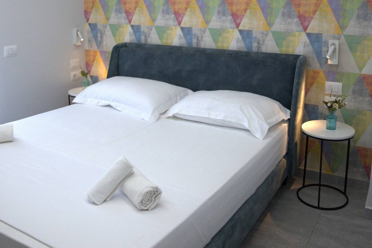 A neatly made bed with white linens and two rolled towels, a gray upholstered headboard, and a colorful geometric-patterned wall.