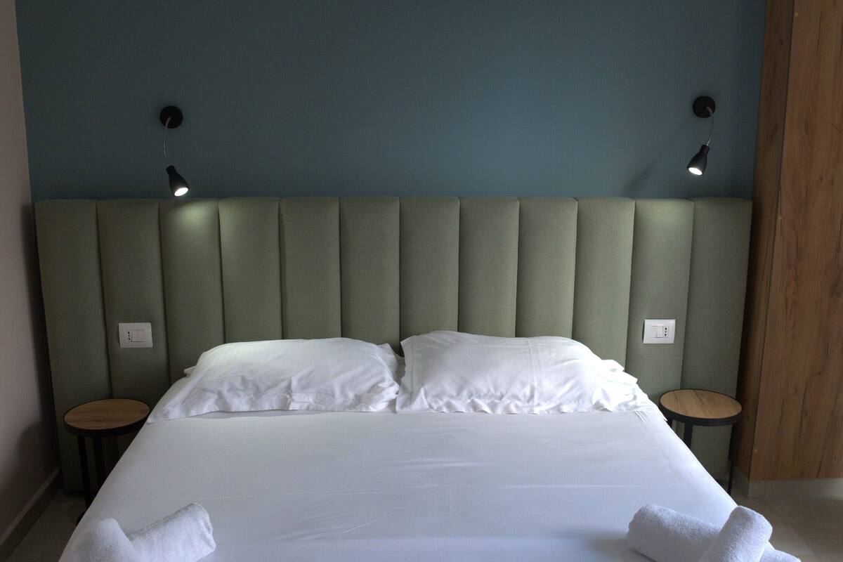 A neatly made bed with white linens and towels, a padded headboard, and two bedside tables with lamps.
