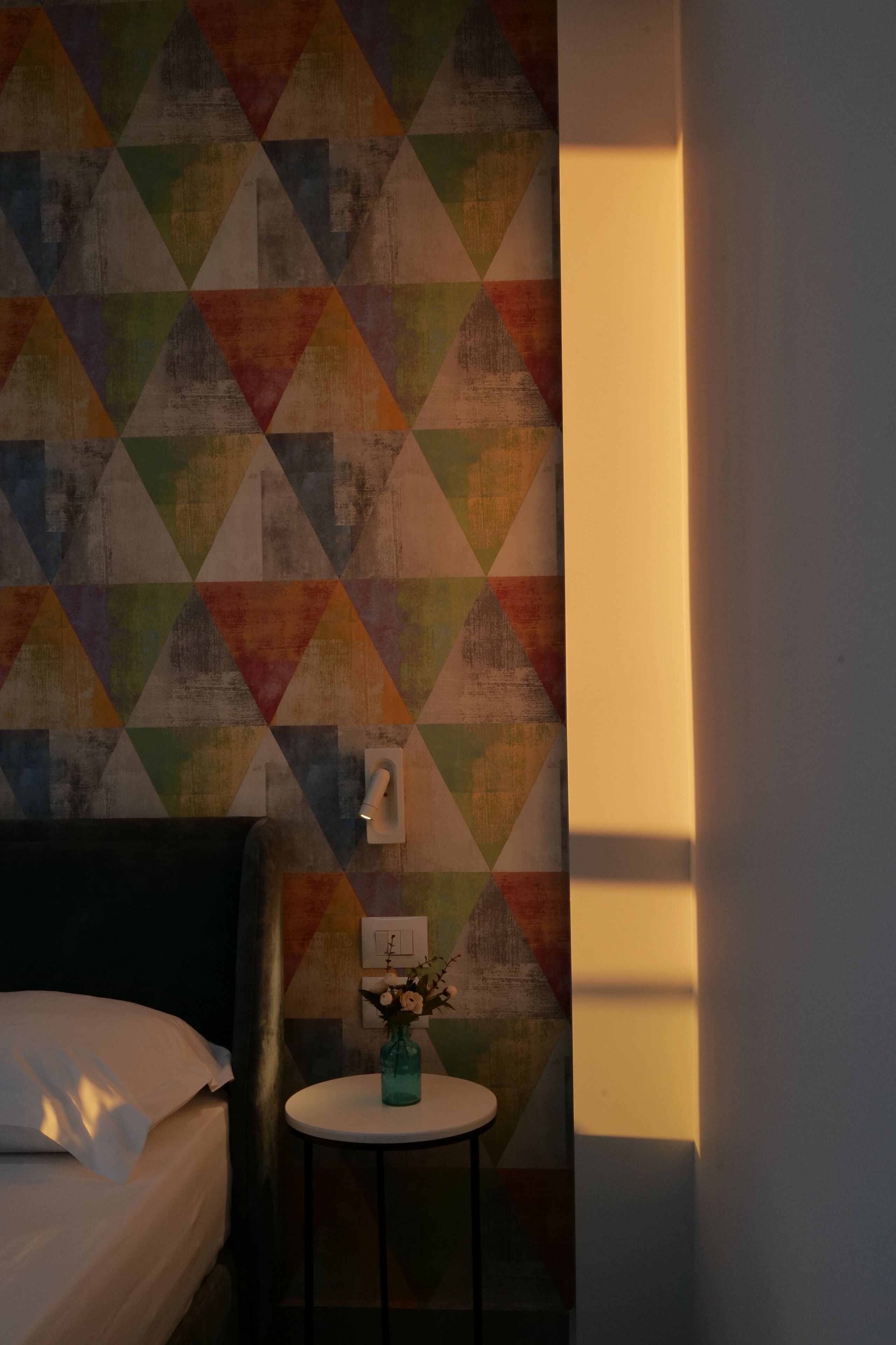 Et værelse med en seng, et lille rundt bord med en vase, og en væg med et farverigt geometrisk mønster.