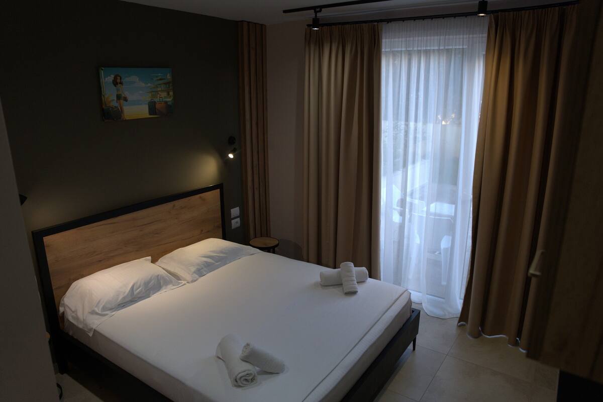 A neatly made bed with white linens and rolled towels, a wooden headboard, a small bedside table, and a painting on the wall.