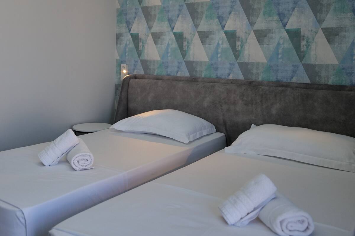 A hotel room with two beds, each with a neatly folded towel. The room features a geometric patterned wall and a modern headboard.