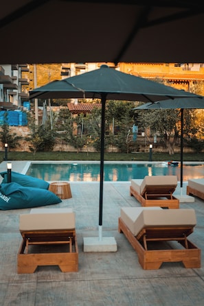 A poolside area with lounge chairs and umbrellas, a pool, and a view of buildings and trees in the background.