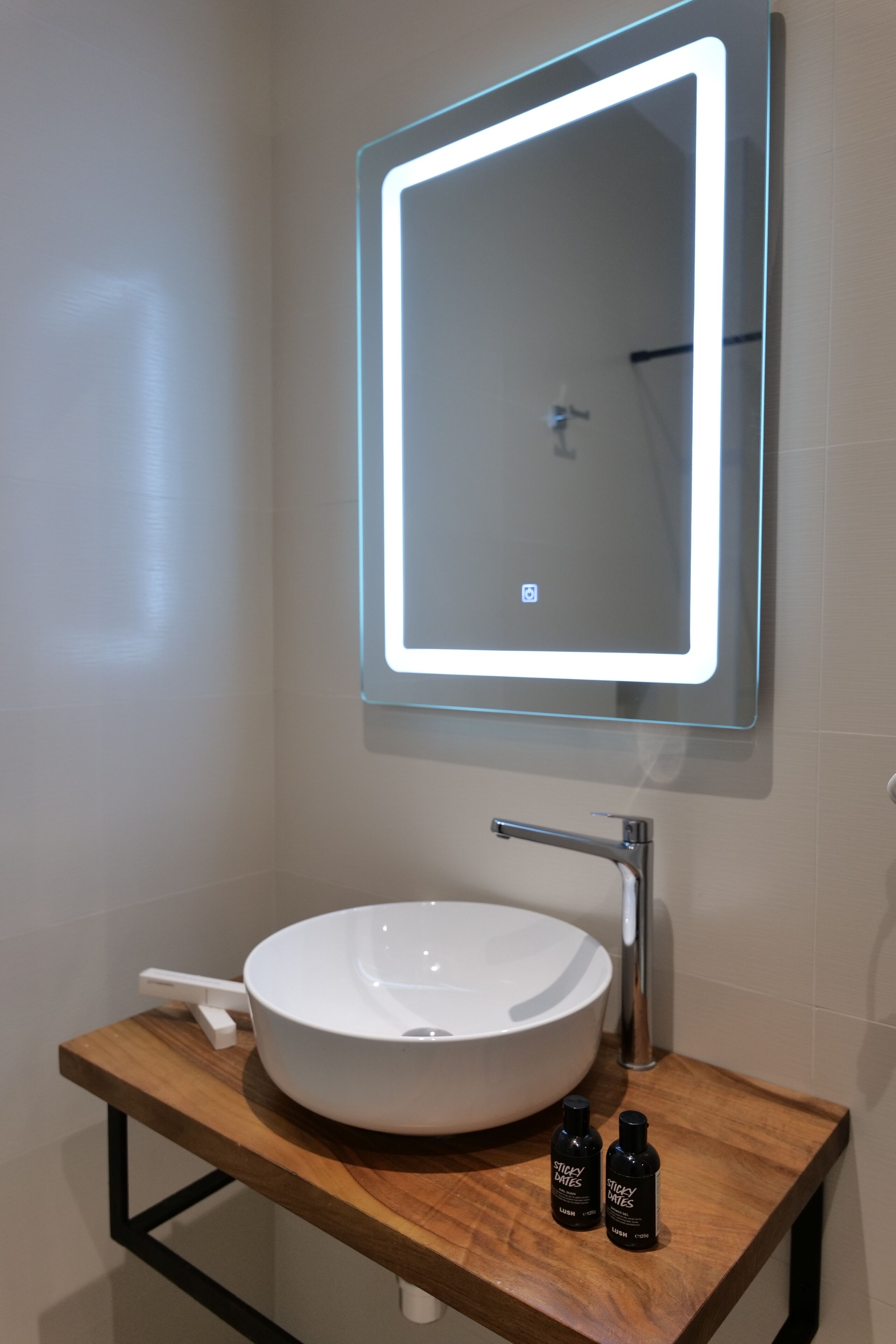 A modern bathroom with a round white sink, a rectangular mirror with LED lighting, and two small bottles on a wooden countertop.