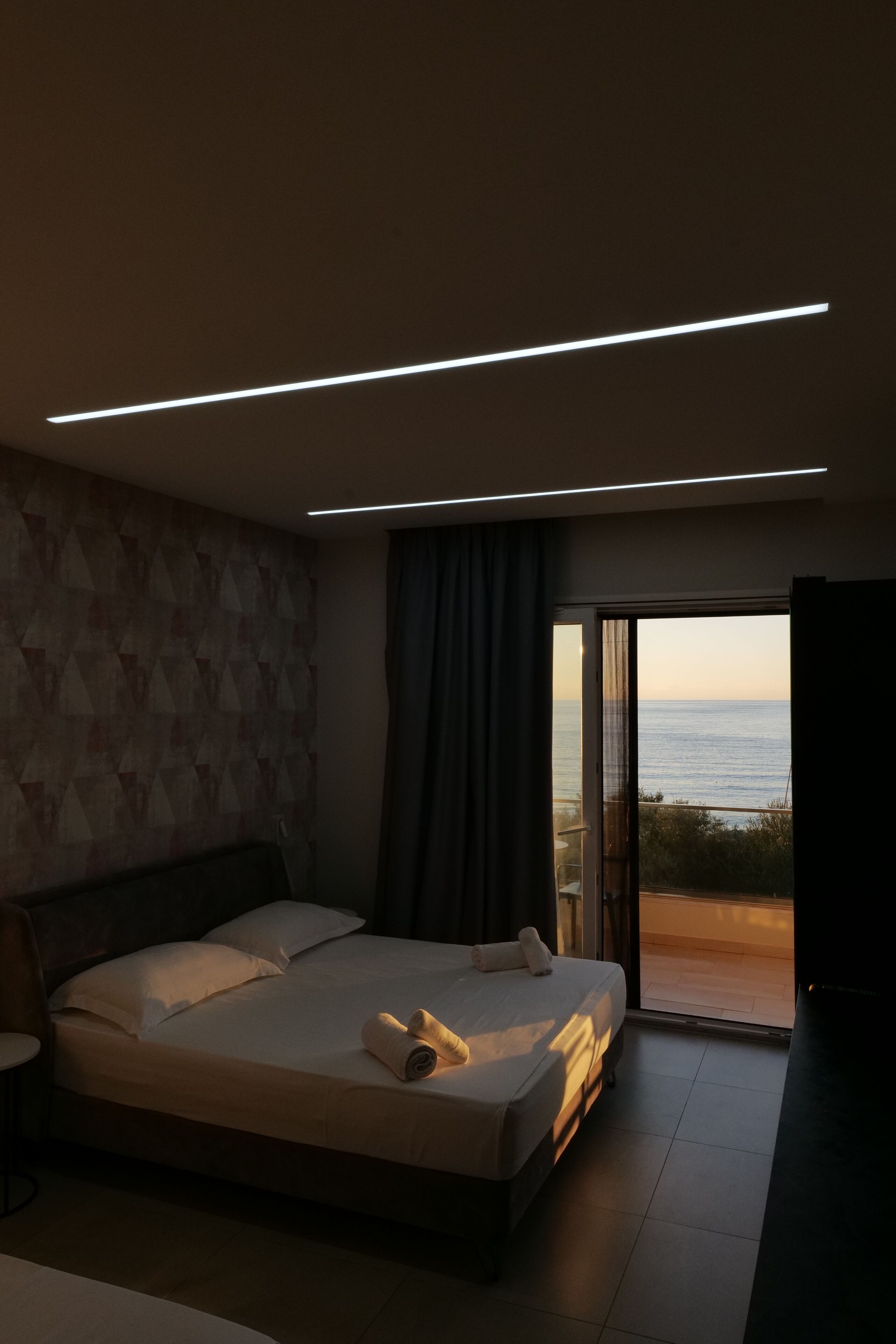A modern bedroom with a large bed, a view of the sea through a sliding door, and a geometric-patterned wall.