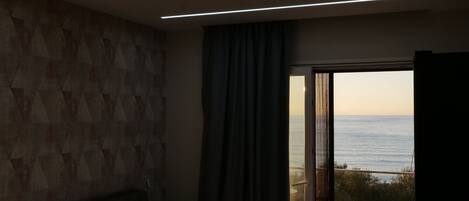 A modern bedroom with a large bed, a view of the sea through a sliding door, and a geometric-patterned wall.