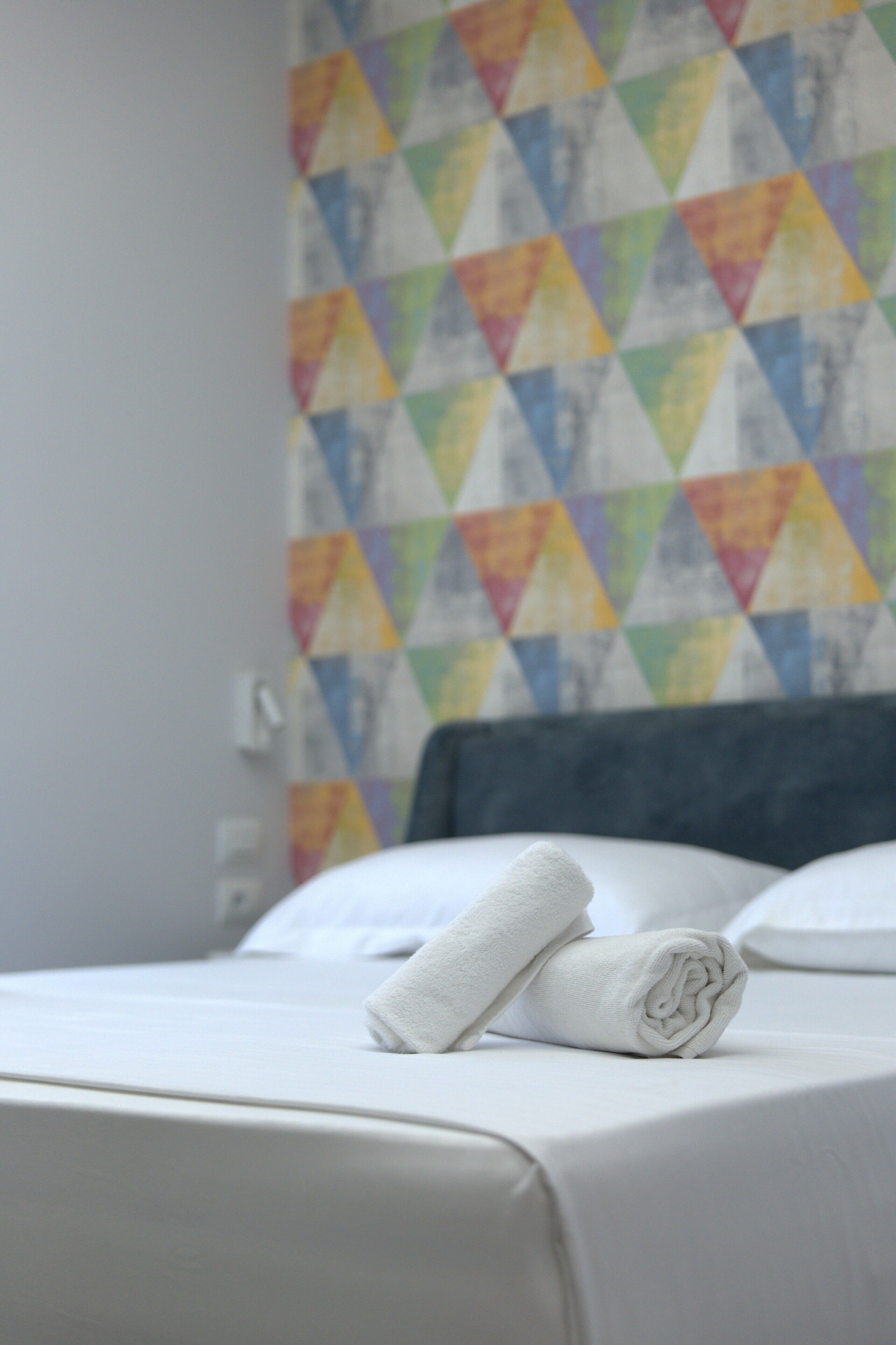 Un lit bien fait, avec une serviette blanche pliée et un oreiller, un mur aux motifs géométriques colorés et une tête de lit sombre.