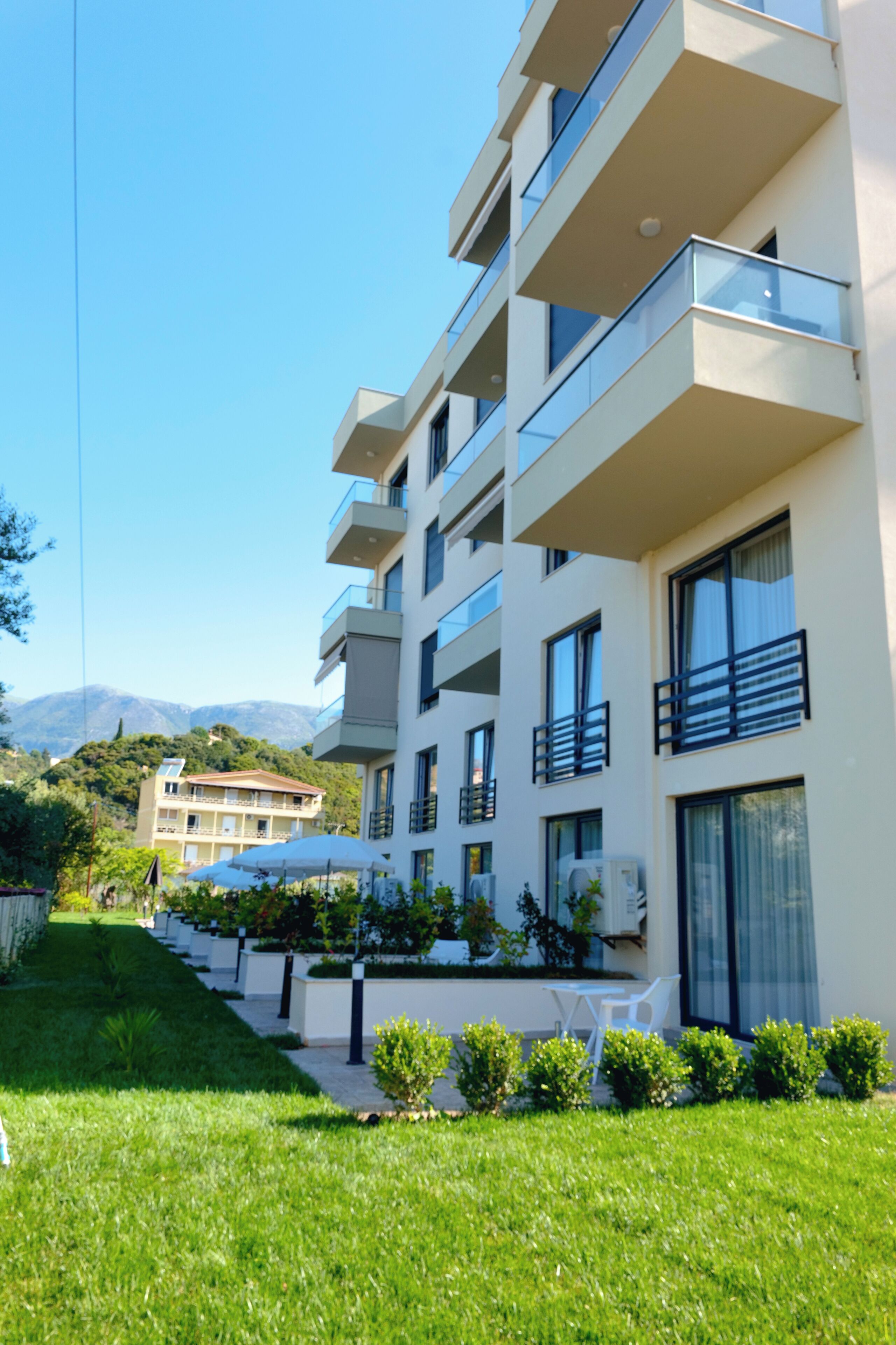 Un hotel de varios pisos con balcones, un jardín con plantas y mobiliario exterior, y montañas al fondo.