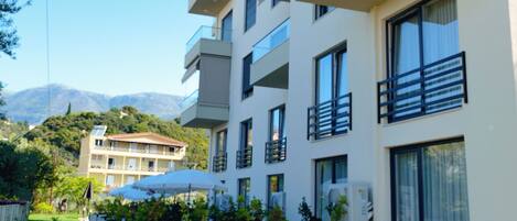 Un hotel de varios pisos con balcones, un jardín con plantas y mobiliario exterior, y montañas al fondo.