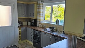 A compact kitchen with a washing machine, a coffee maker, and a window with a view of trees. - 2 bedroom chalet bungalow, short drive to The New Forest and Bournemouth beaches (Poole)