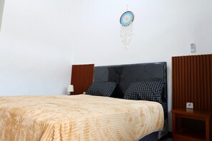 A neatly made bed with a patterned blanket, a wall-mounted dreamcatcher, and a bedside table with a lamp. - Pondok Dete Guesthouse (Denpasar)