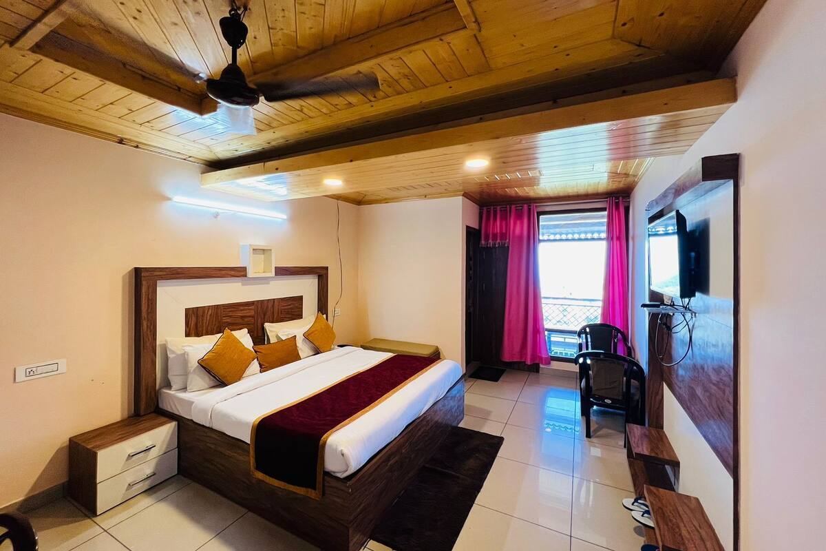 Ein Hotelzimmer mit Holzbalkendecke, einem Bett mit weißen Leinen und einer roten Tagesdecke, einem Schreibtisch mit Stuhl und einem an der Wand befestigten Fernseher.