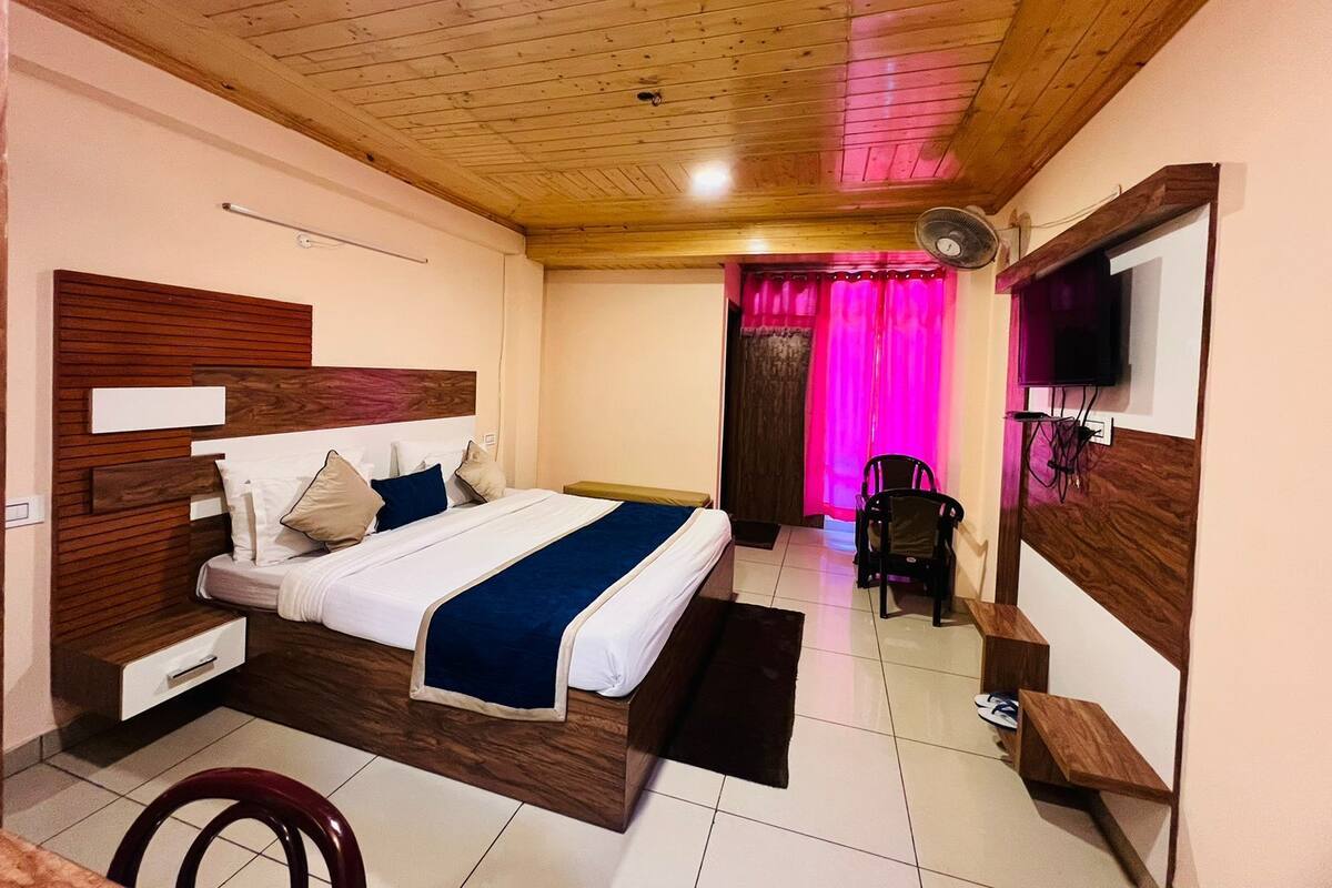 Ein Schlafzimmer mit Holzbalkendecke, einem Bett mit blauer Bettdecke, einem an der Wand montierten Fernseher und einem Sessel in der Nähe des Fensters.