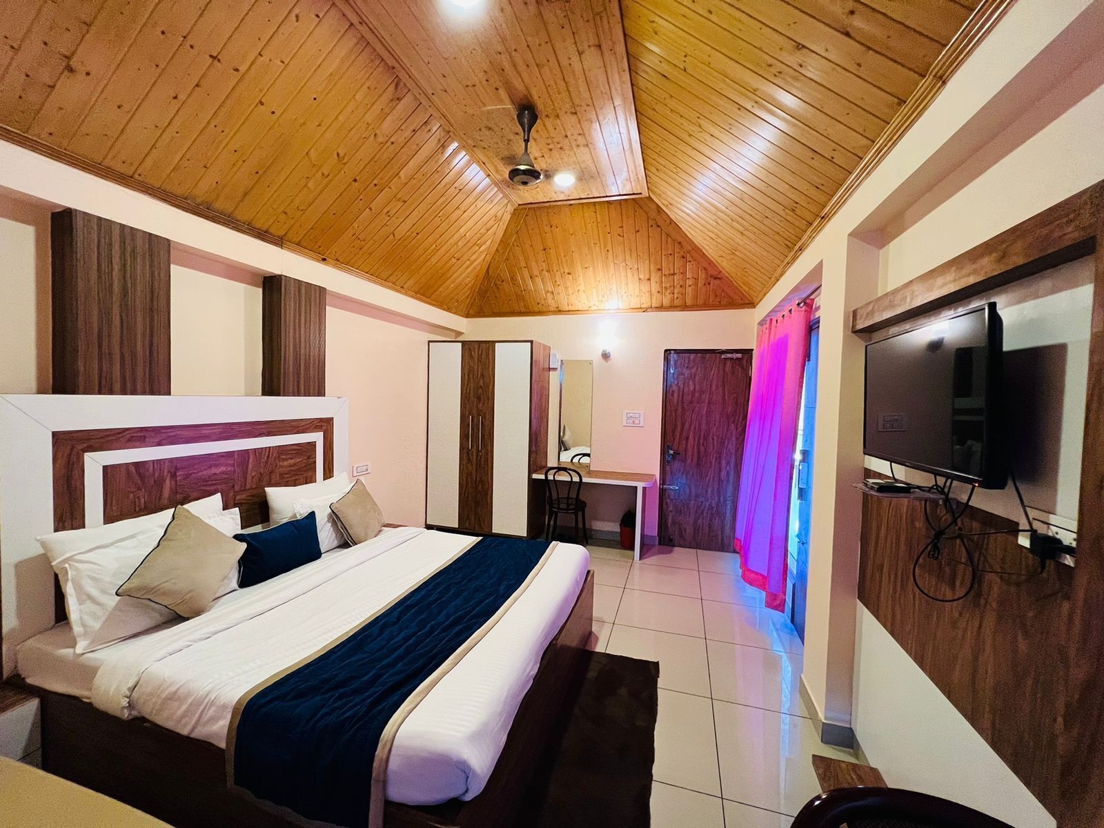 Une chambre avec un plafond en bois, un lit avec une tête de lit, un bureau, une chaise, une armoire et un téléviseur fixé au mur.