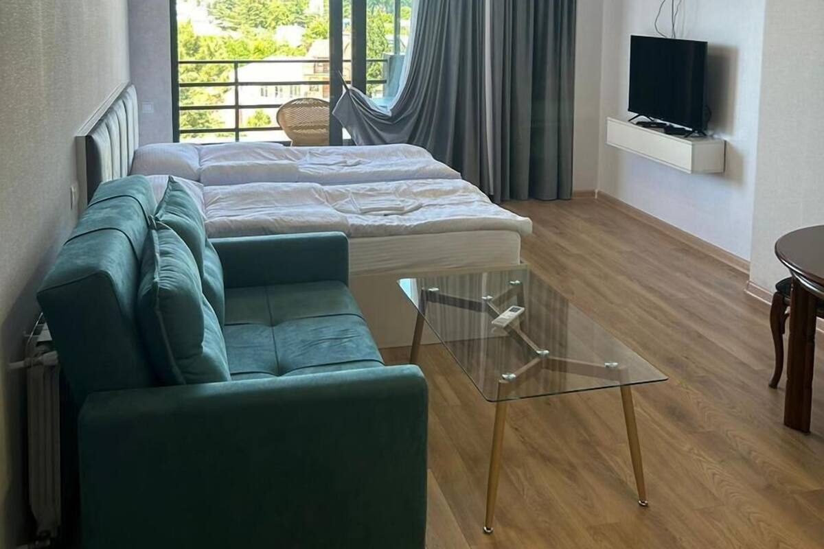 A modern hotel room with a bed, a sofa, a glass table, a TV, and a balcony with a view of greenery.