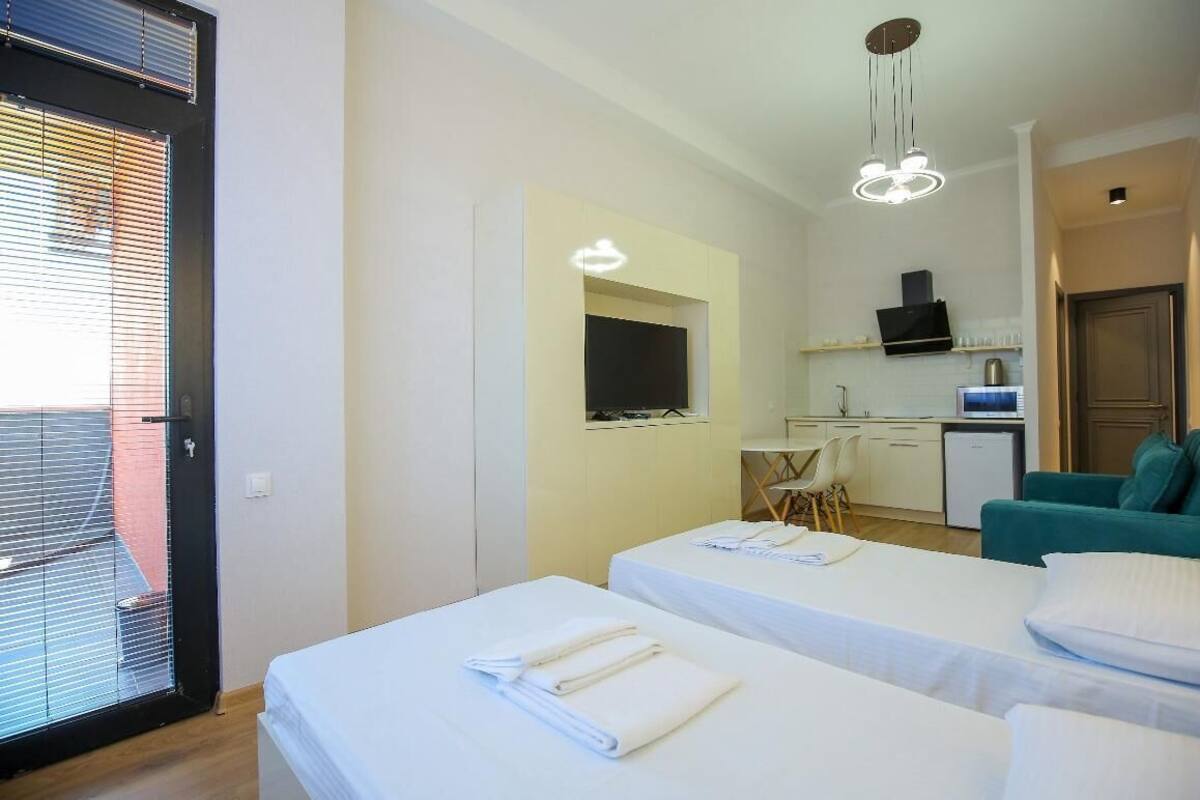 A hotel room with two beds, a kitchenette, a TV, and a balcony with blinds.