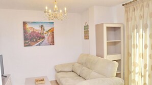 A living room with a beige sofa, a small white table, a chandelier, a painting, a cabinet, and a polka dot curtain. - Apartamento El Mirador (Conil de la Fra.)