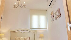 A bedroom with a bed, two bedside lamps, a chandelier, a window with blinds, and two wall-mounted pictures. - Apartamento El Mirador (Conil de la Fra.)