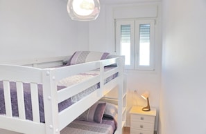 Un letto a castello con una finestra sopra, un comodino con una lampada e un piccolo mobiletto contenitore.