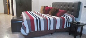 Una cama con colcha a rayas, cabecera capitonada, mesita de noche y un armario de color oscuro.