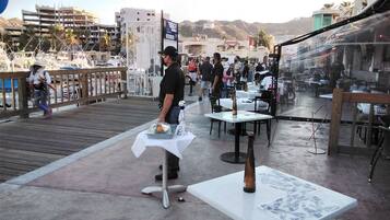 Un área de comedor al aire libre con mesas y sillas, un camarero, y vistas a edificios y colinas al fondo.