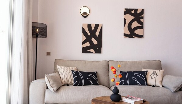 Uma sala de estar moderna com sofá, almofadas decorativas, obras de arte na parede, abajur e um vaso com flores sobre uma mesa lateral.