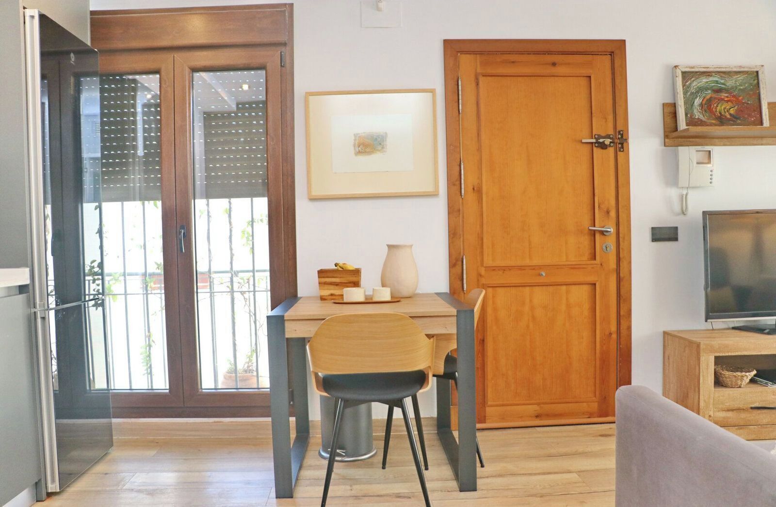 A room with a wooden door, a framed picture on the wall, a table with chairs, and a television on a wooden stand.