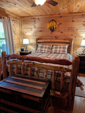 A wooden cabin bedroom with a bed, two bedside tables, a lamp, and a decorative wall hanging. - Cozy Rustic Cottage w/Hot Tub & Fire Pit near Lake (Ellijay)