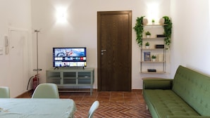 Una sala de estar con televisor, un sofá verde, una mesa de comedor con sillas y un mueble pequeño.