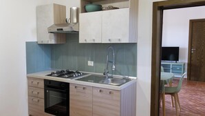 Cocina compacta con estufa a gas, lavamanos y horno. Hay un mueble de madera sobre la estufa y una pequeña estantería con un cuenco. Se ve una silla en la habitación contigua.