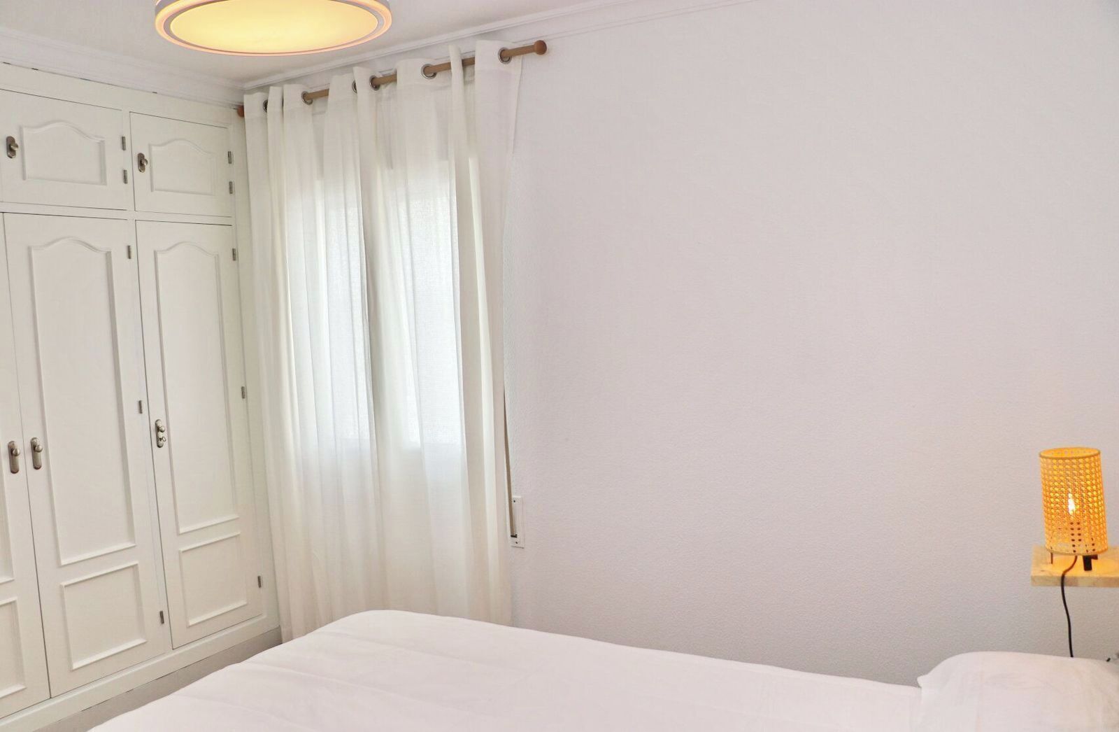 A bedroom with a bed, white curtains, a wardrobe, and a wall-mounted lamp.