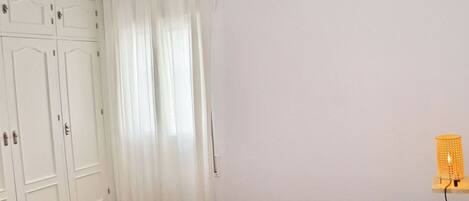 A bedroom with a bed, white curtains, a wardrobe, and a wall-mounted lamp.