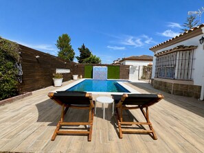 Una piscina con dos sillas de descanso, una terraza de madera y una pared blanca con una ventana.